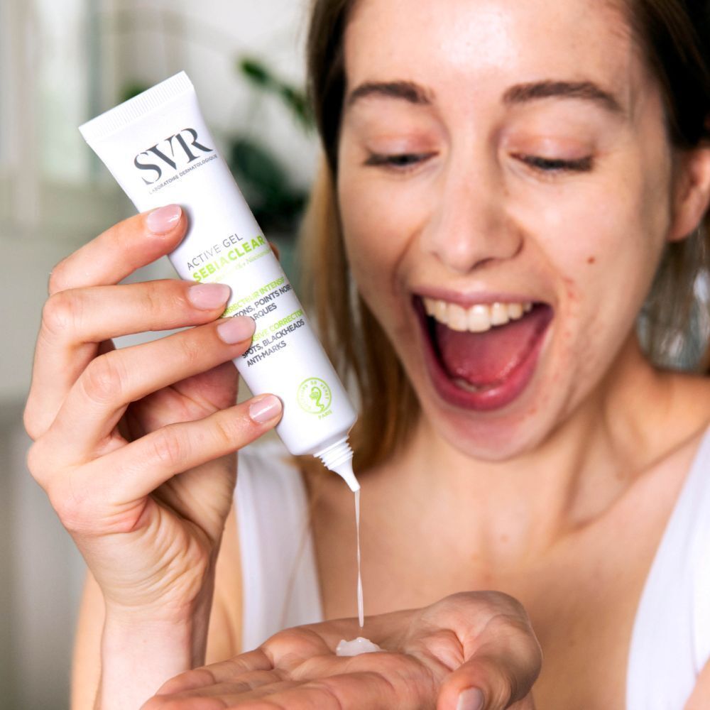 Une femme tient un tube de SVR SEBIACLEAR Active Gel. Le produit est pressé sur la main. La femme sourit.