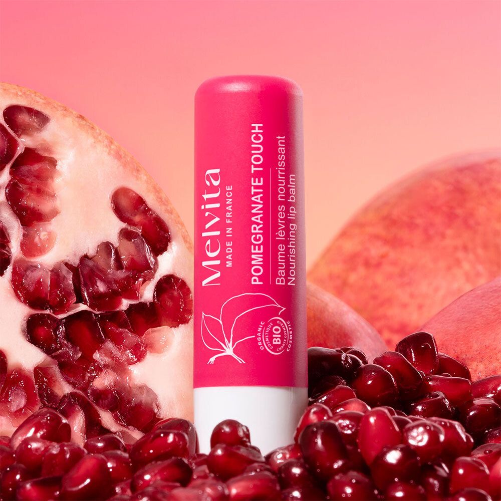 Rosa Lippenbalsam-Stift mit Melvita-Logo, Granatapfel-Motiv und Bio-Siegel. Umgeben von Granatapfel-Früchten und -Kernen.