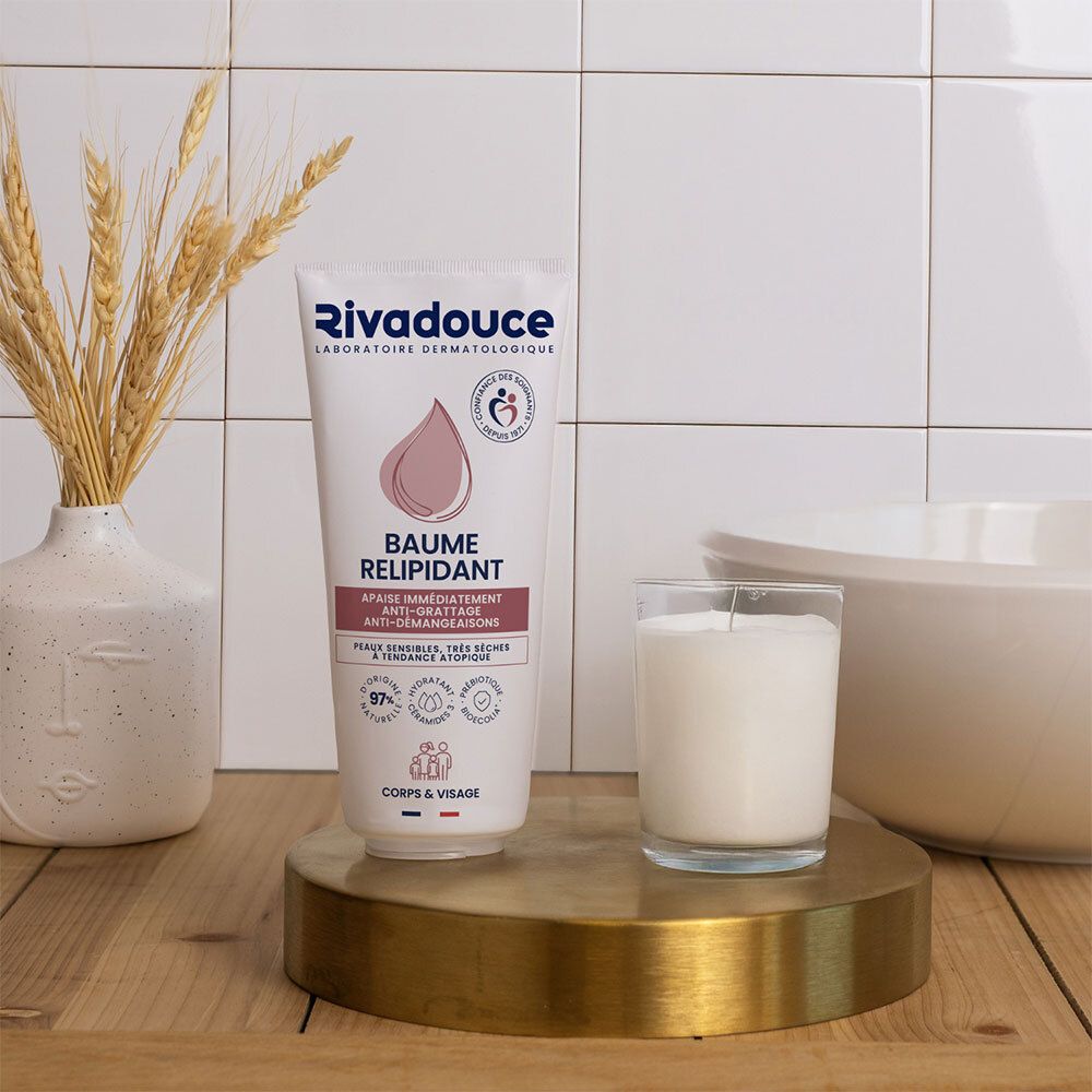 Produkt auf goldener Oberfläche. Weisse Tube mit Rivadouce-Logo. Kerze und Vase mit Getreideähren im Hintergrund. Fliesenwand.