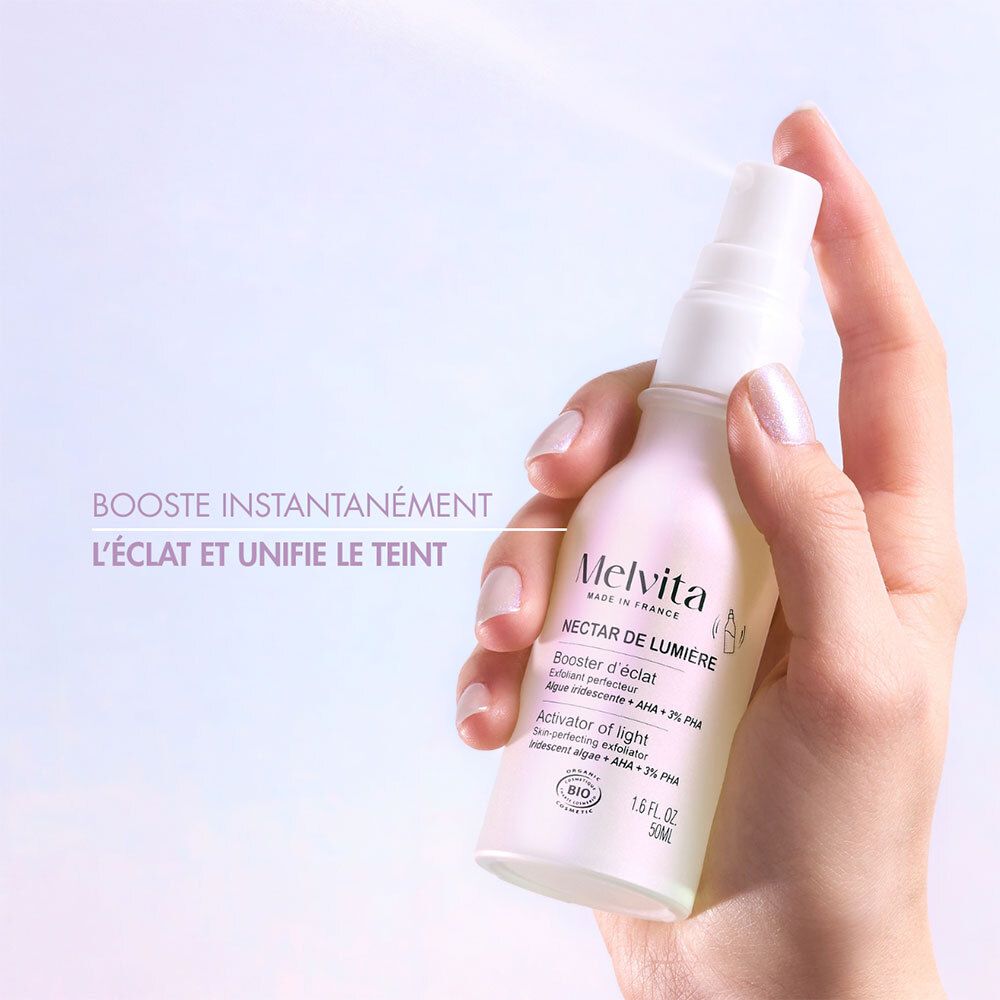 Hand hält Flasche mit Zerstäuber. Text: Booster instantanément l'éclat et unifie le teint. Melvita, Nectar de Lumière.