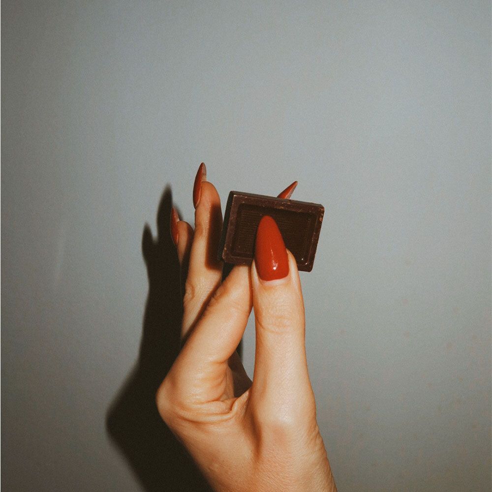 Hand mit rotem Nagellack hält ein einzelnes Stück dunkle Schokolade.