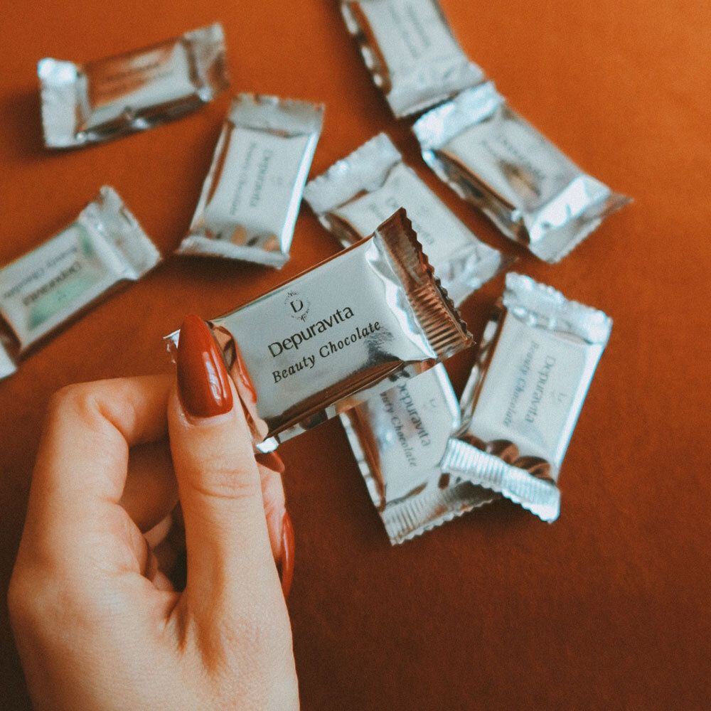 Hand mit rotem Nagellack hält eine einzelne verpackte Depuravita Beauty Chocolate.
