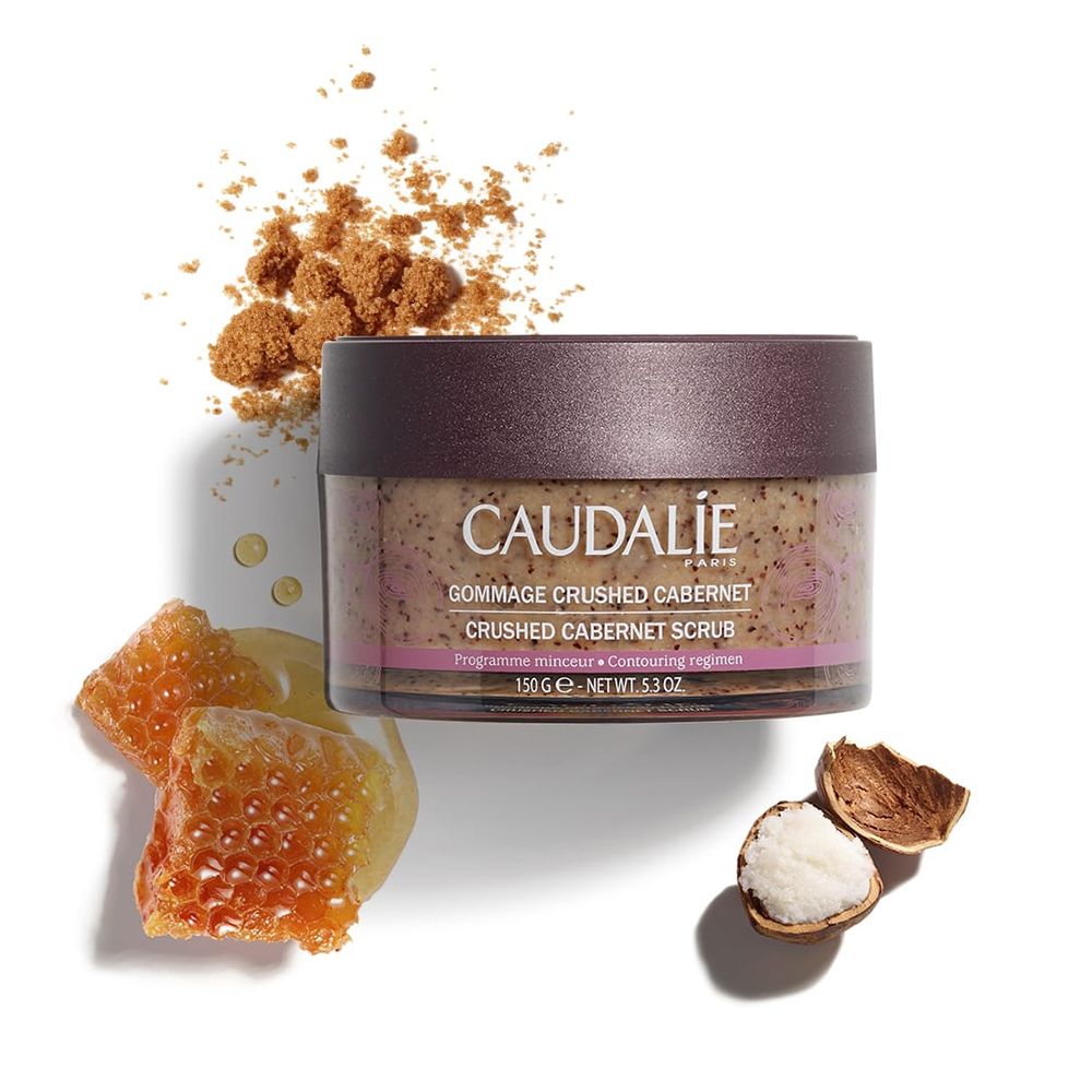Pot de gommage et ingrédients. Couvercle marron foncé. Inscription: CAUDALIE, Gommage Crushed Cabernet, Crushed Cabernet Scrub. 150g.
