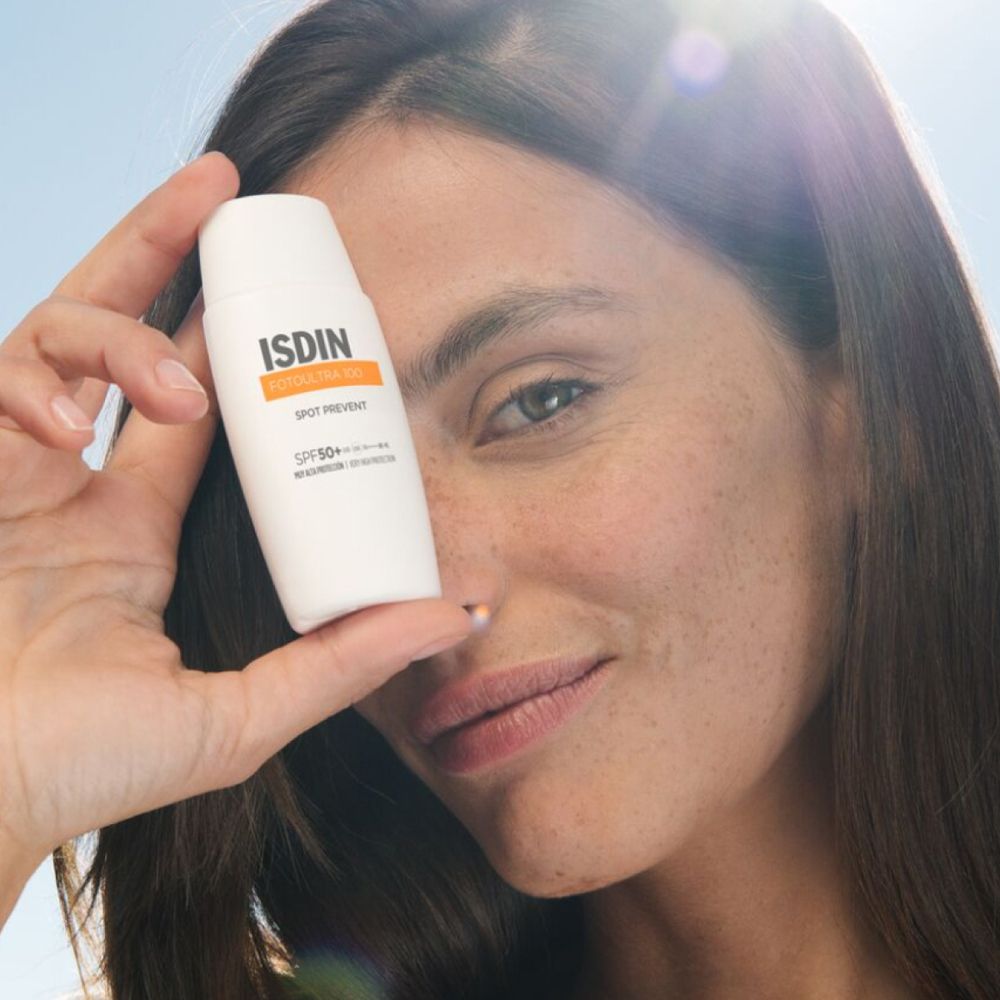 Femme tenant un flacon blanc avec le logo ISDIN devant son visage. Nom du produit : ISDIN FOTOULTRA 100 Spot Prevent. Peau avec des taches de rousseur.