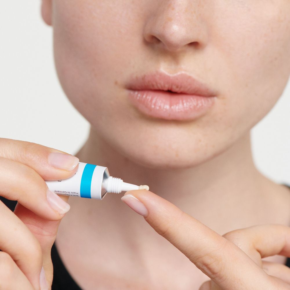 Nahaufnahme einer Person, die Creme aus einer Tube auf den Finger aufträgt. Lippen im Hintergrund.
