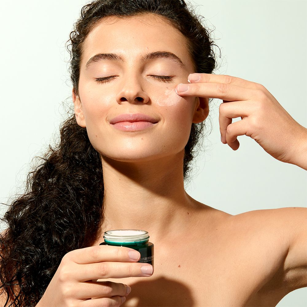 Frau mit geschlossenen Augen, berührt Gesicht mit Finger. Kleine Creme-Dose sichtbar. Keine weiteren Details.