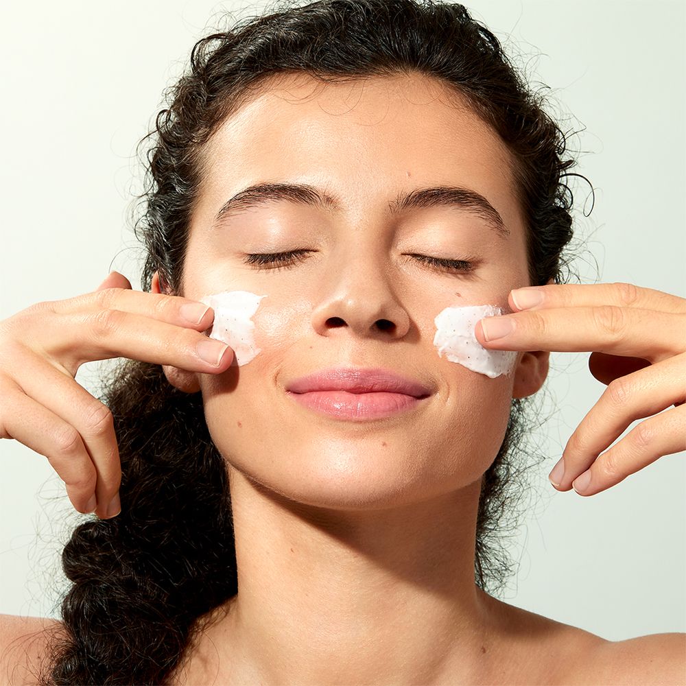 Frau mit geschlossenen Augen, trägt Produkt auf Wangen. Produkt ist weiß. Hände berühren Wangen.