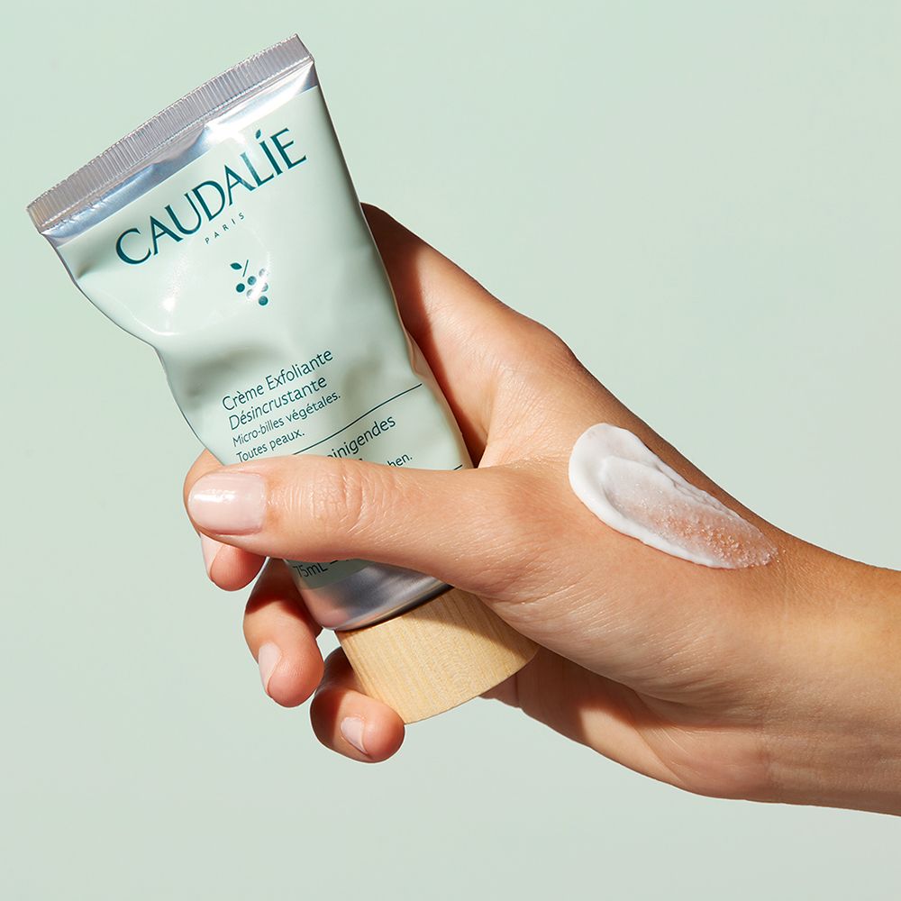 Main tenant un tube de gommage facial CAUDALÍE. Goutte de gommage sur la main. Bouchon en bois.