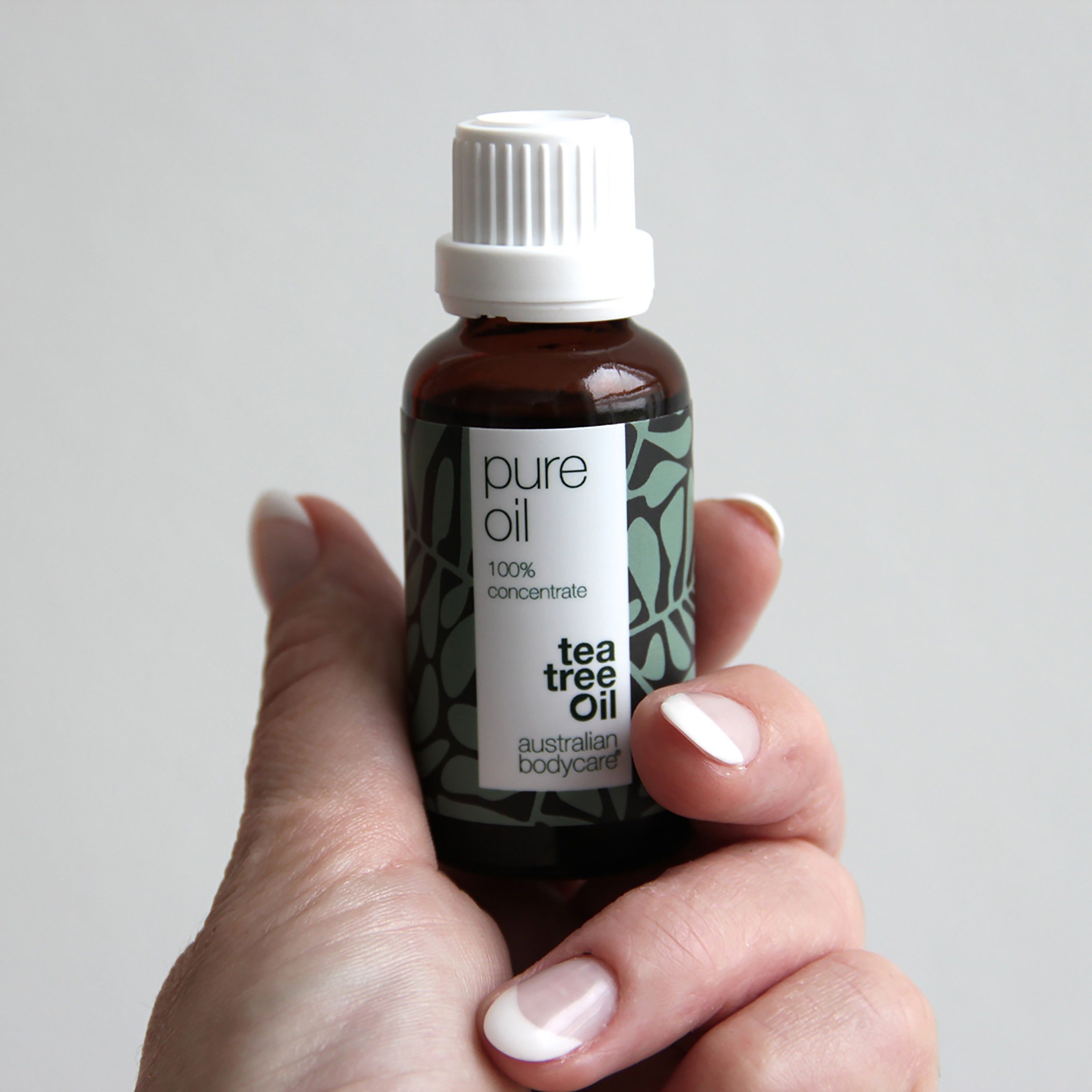 Flacon en verre brun avec bouchon blanc, tenu par une main. Étiquette avec texte : Pure Oil, 100% concentrate, Tea Tree Oil.