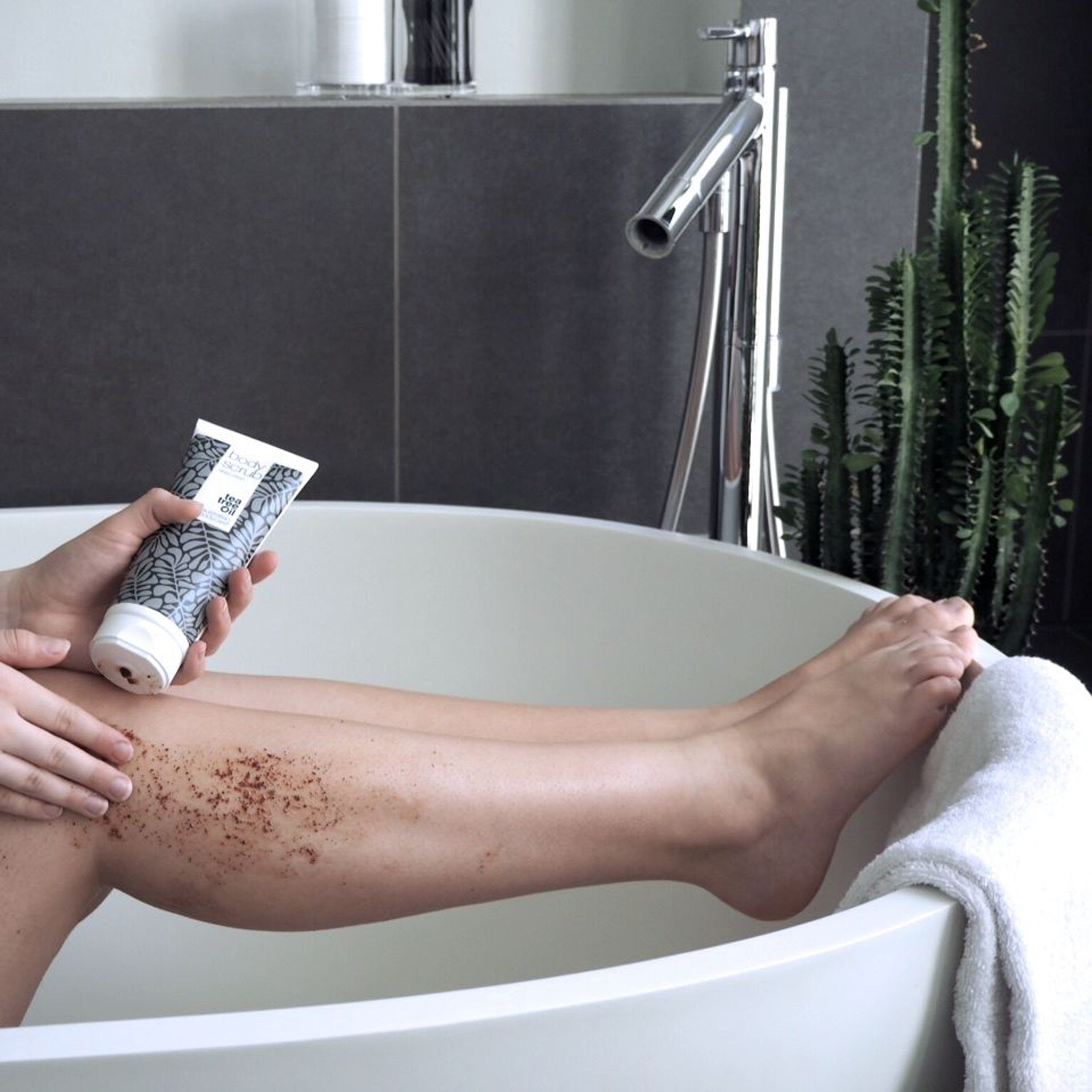 Personne tenant un tube « Body Scrub ». Jambe dans une baignoire. Marque : Australian Bodycare.
