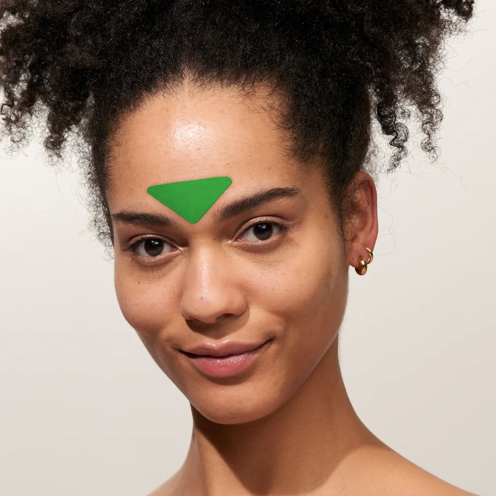 Femme avec patch triangulaire vert sur le front. Boucle d'oreille dorée. Peau claire, cheveux foncés.