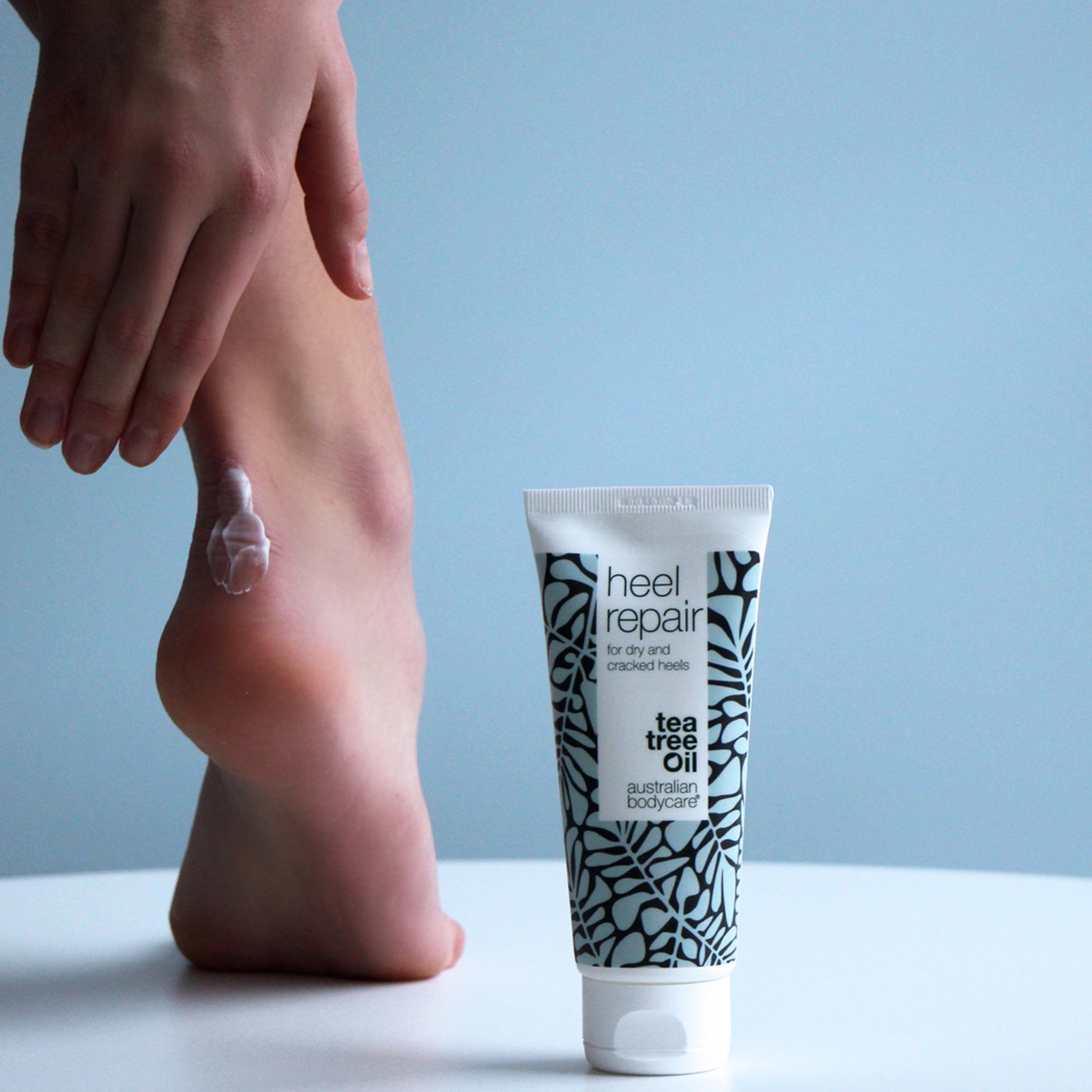 Tube de crème pour talons. Blanc, floral, texte: Heel Repair, pour talons secs et crevassés. Personne applique crème sur talon.