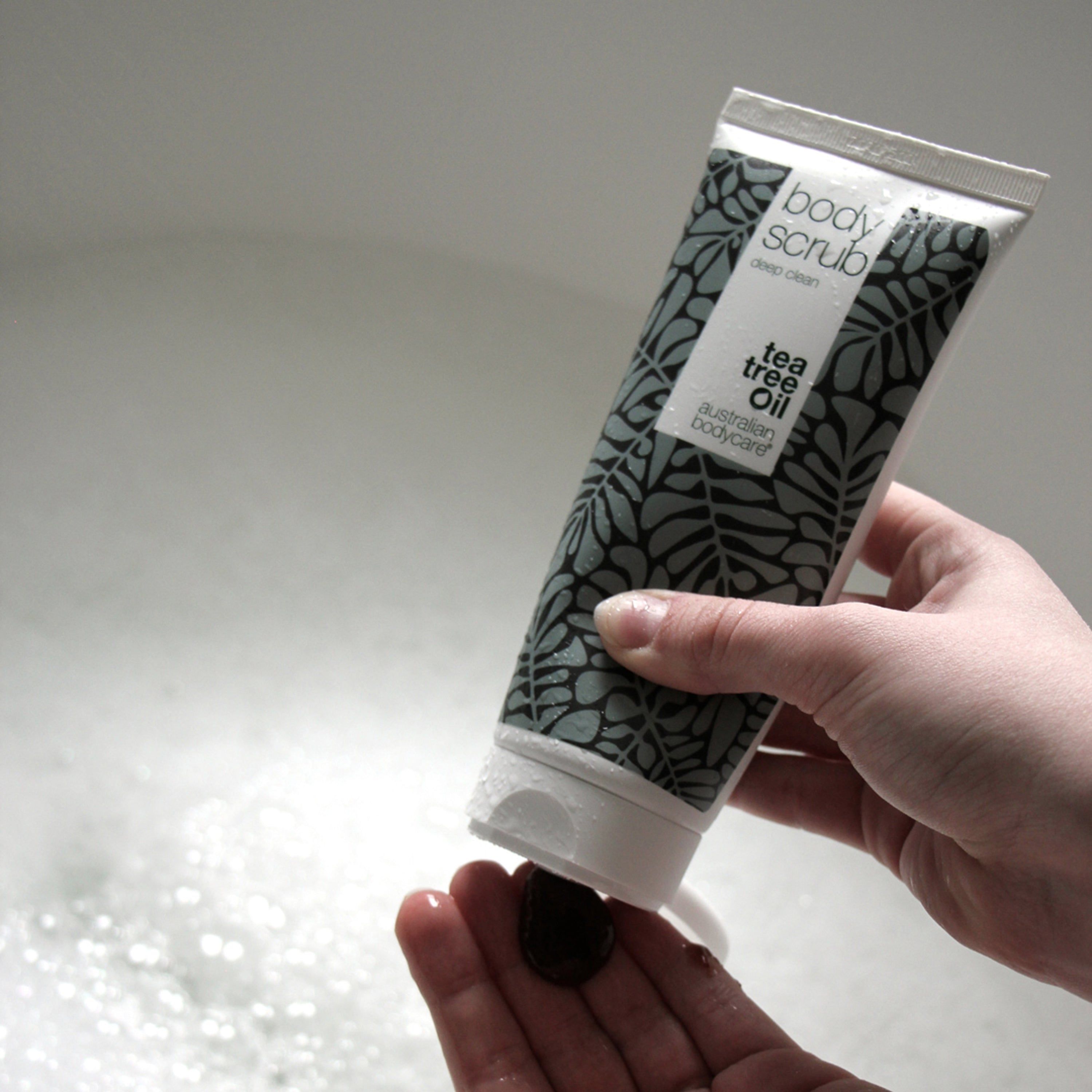 Personne pressant du gommage d'un tube. Marque : Australian Bodycare. Texte : Body Scrub. Tea Tree Oil.
