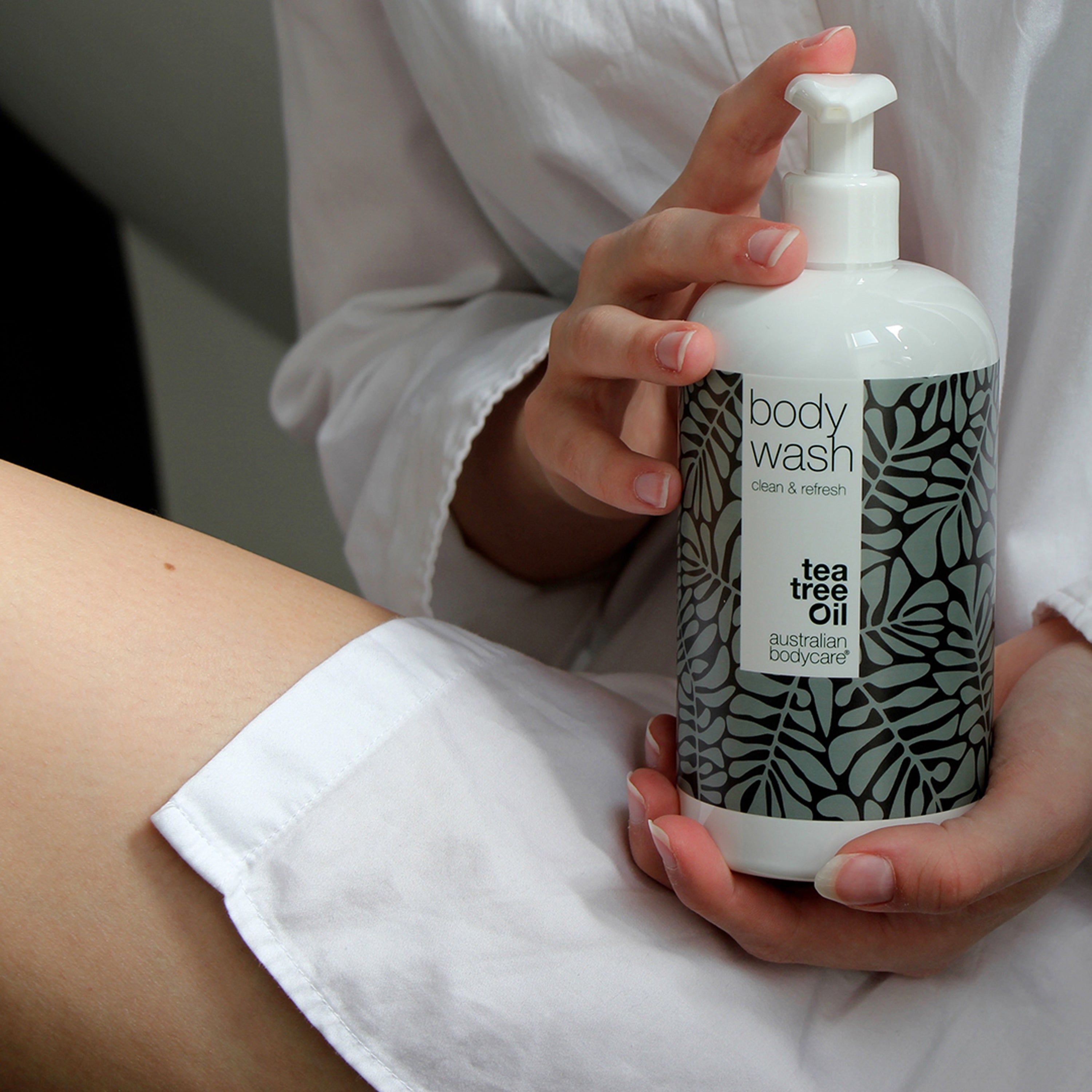 Personne tenant un flacon blanc « body wash » avec motif feuilles. Texte « Tea Tree Oil ».