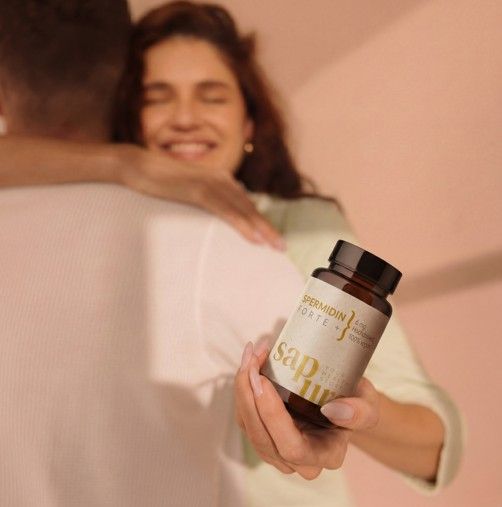 Personne tenant un flacon brun de capsules de spermidine. Inscription : Spermidine Forte +, 100% végétalien. Logo SAPURA. La personne embrasse une autre.