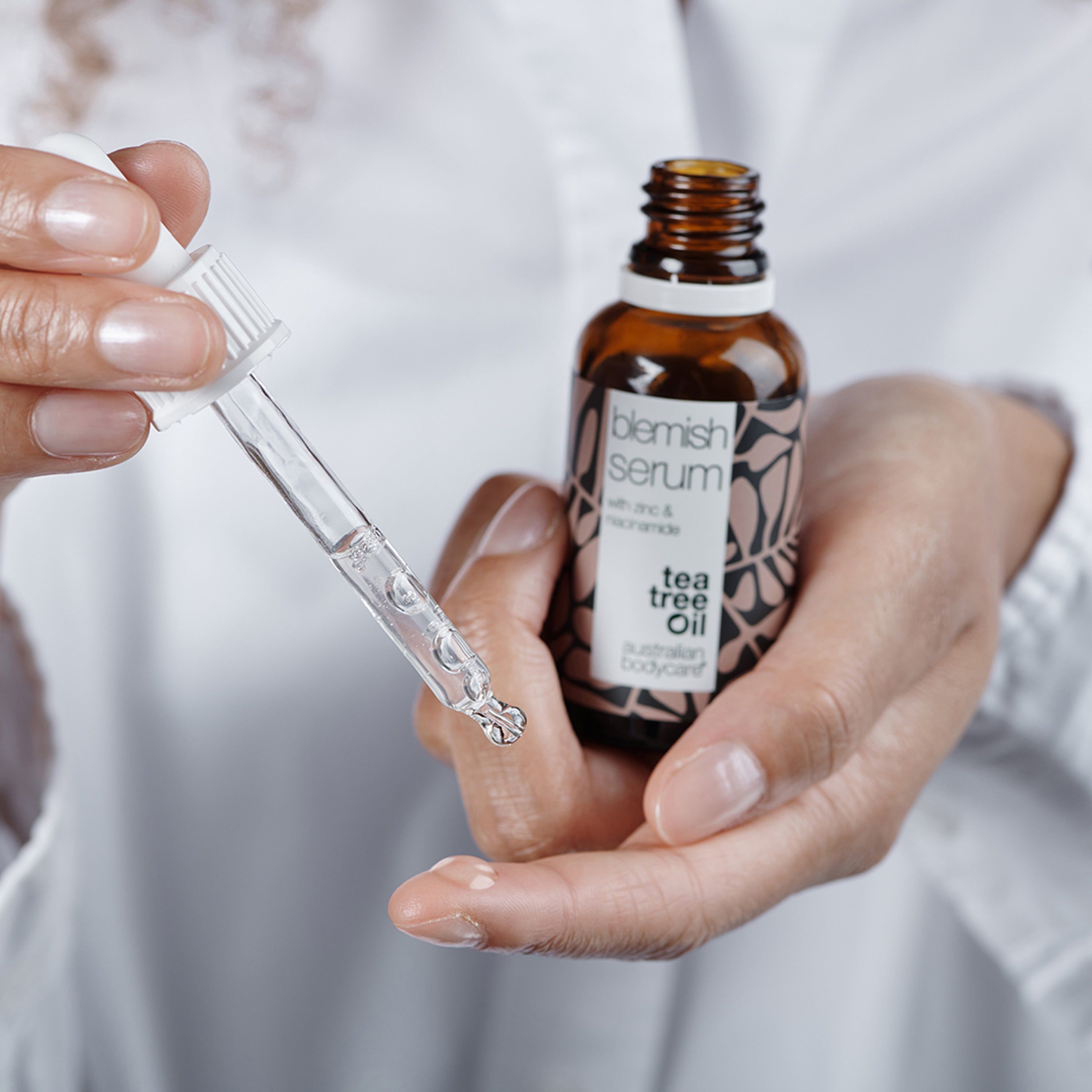 Mains tenant une pipette avec liquide et un flacon de Blemish Serum. Inscription : Blemish Serum, Tea Tree Oil. Marque : Australian Bodycare.