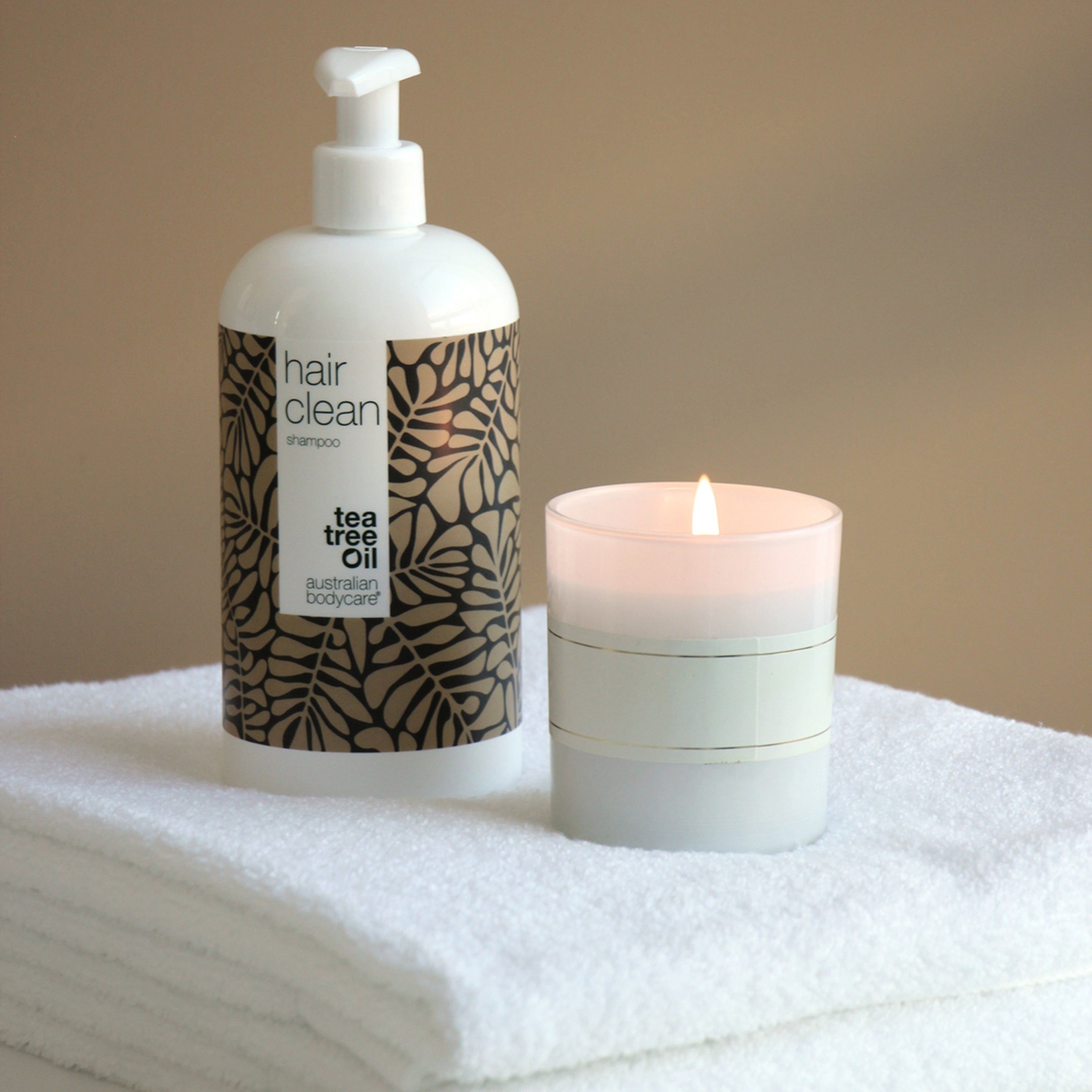 Flacon de shampooing et bougie sur serviettes. Récipient blanc avec motif de feuilles marron. Inscription : hair clean, australian bodycare.