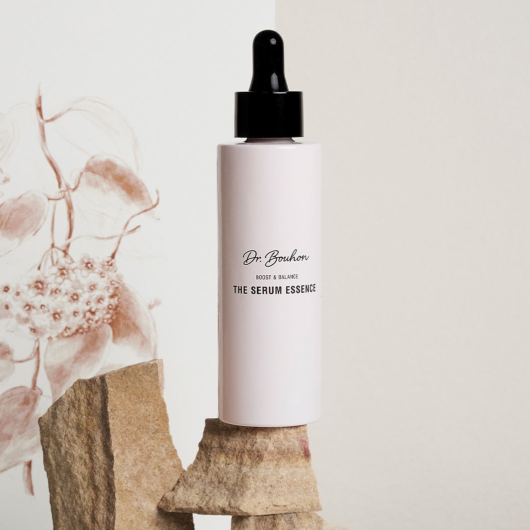 Rosa Flasche mit schwarzem Deckel auf Steinen. Aufschrift: Dr. Bouhon, THE SERUM ESSENCE. Hintergrund: floral.